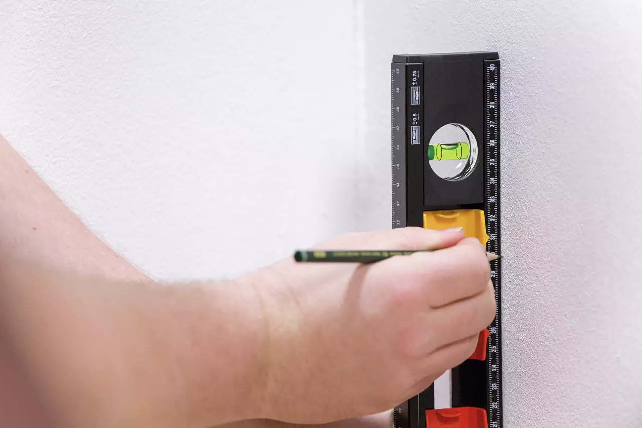 A person using a black and yellow level on a white wall, marking with a pencil.