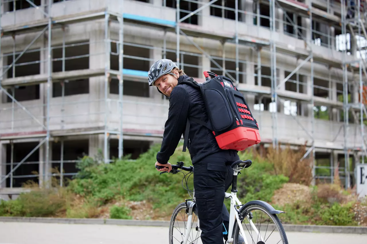 A person on a bicycle wearing a helmet and a large black and red backpack in front of a building under construction.
