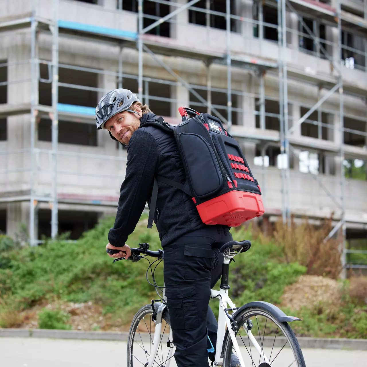 Eine Person auf einem Fahrrad mit Helm und großem schwarz-rotem Rucksack vor einem Rohbau.