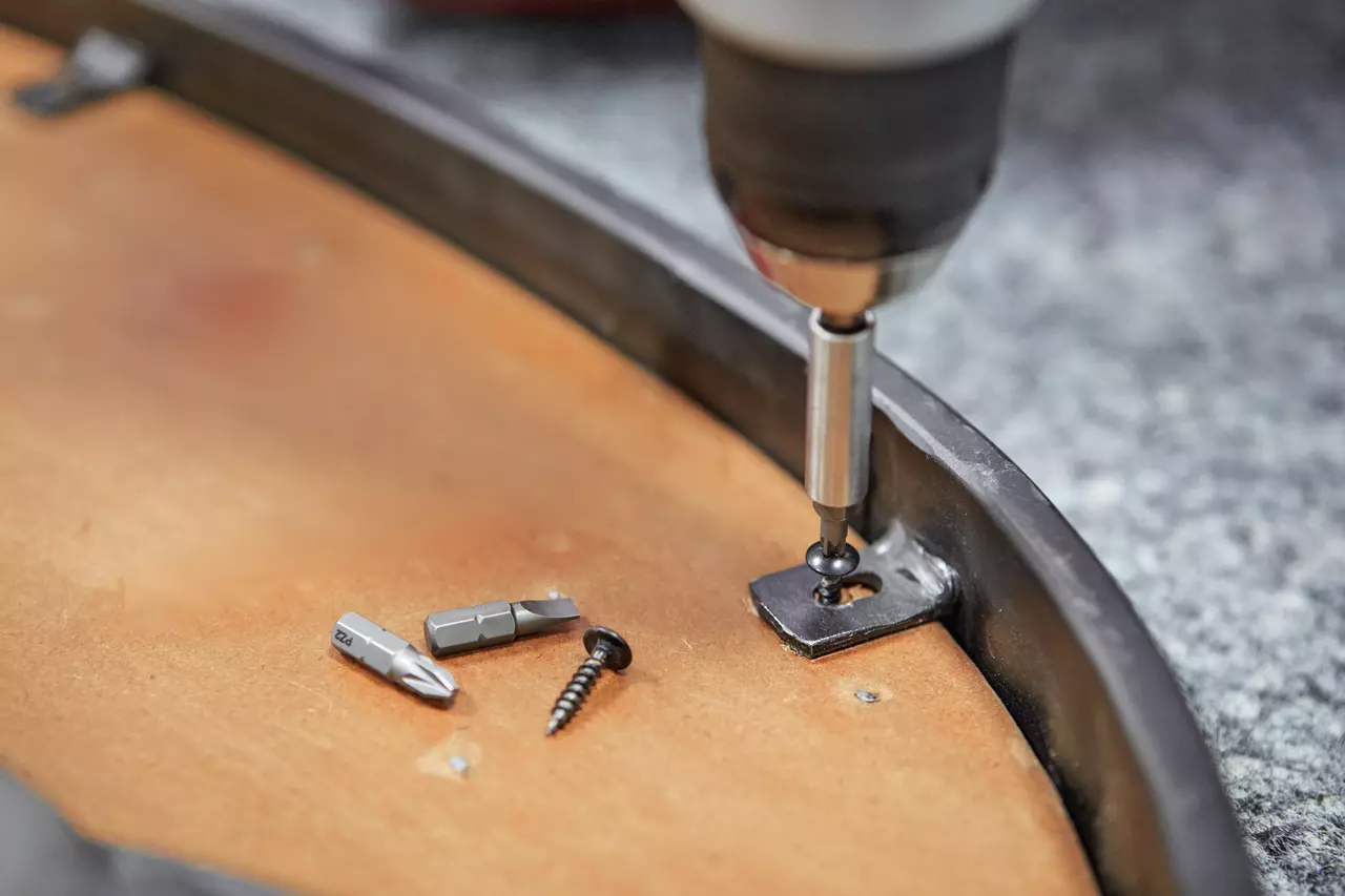 Electric screwdriver screwing a black screw into a metal bracket on wood, with bits and screw nearby.