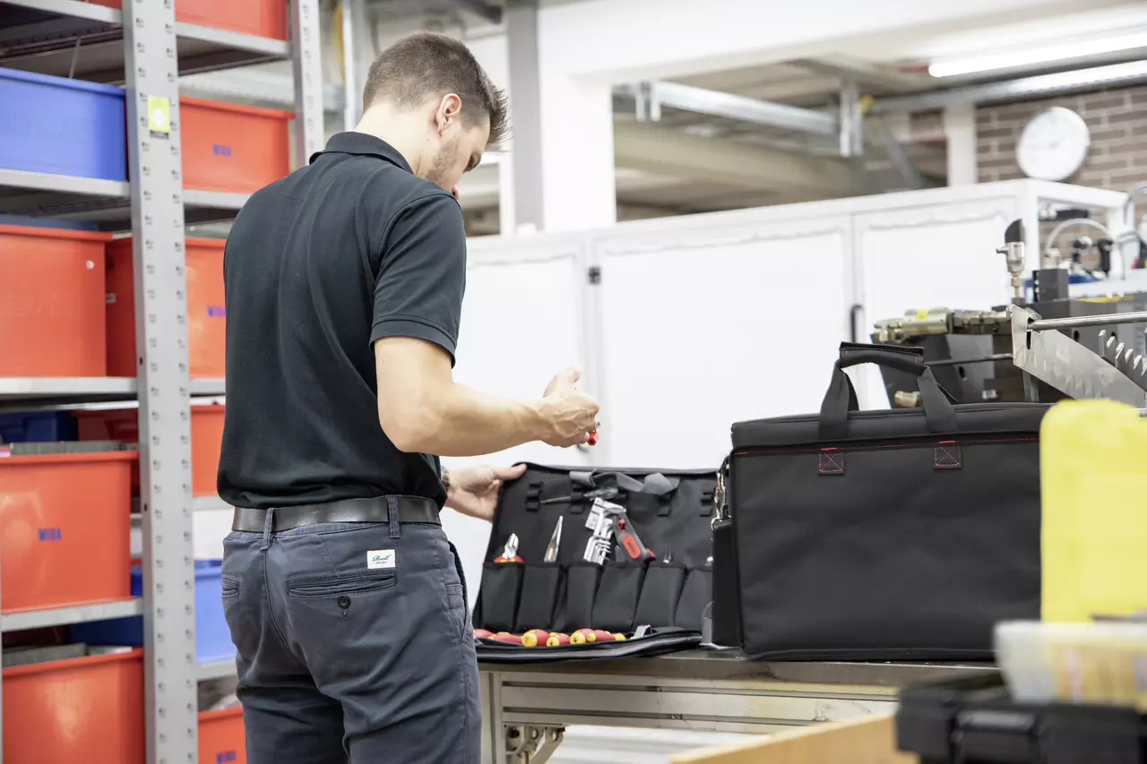 A person in a workshop is organizing tools in a black tool bag with red containers on shelves nearby.