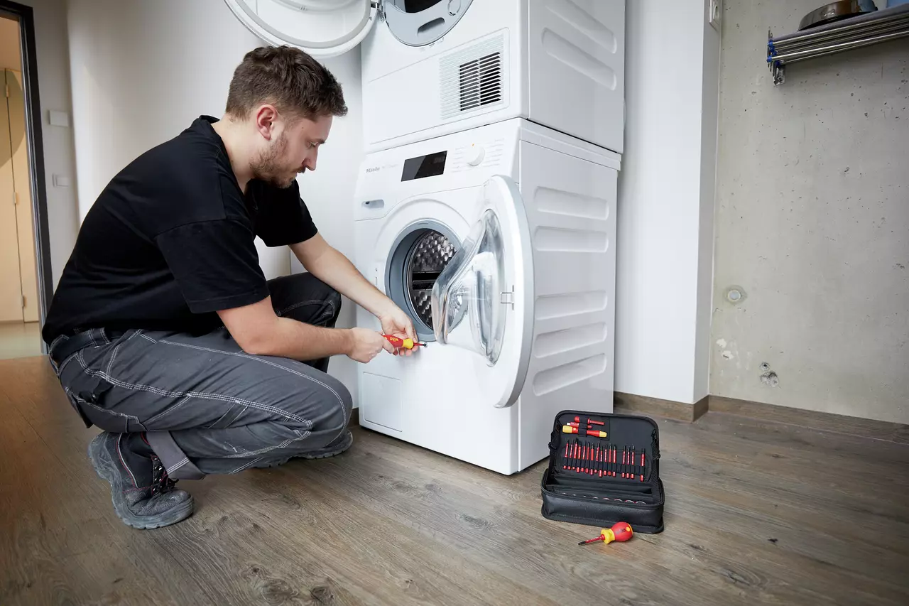 A person uses a screwdriver to repair a washing machine next to a toolset on the floor.