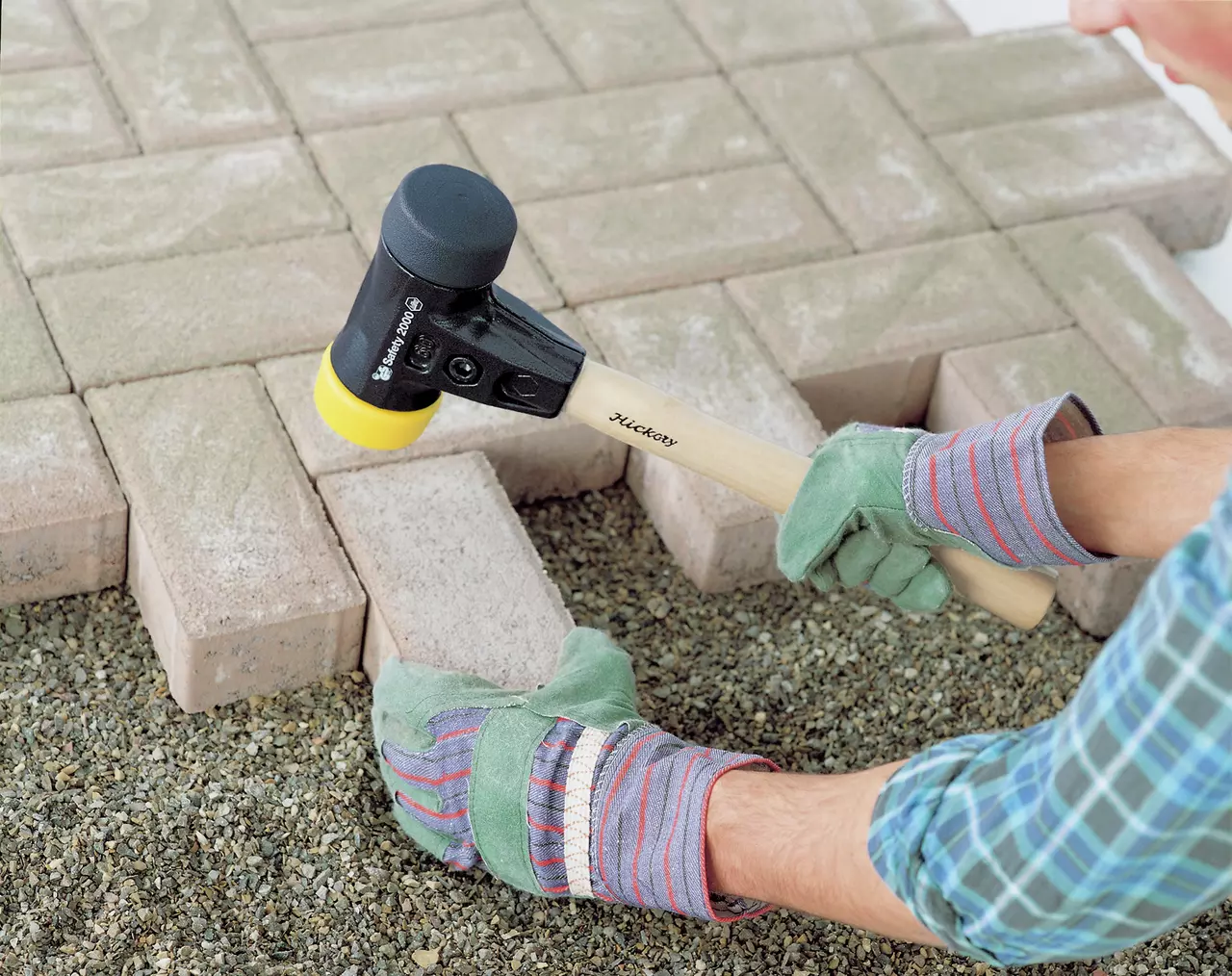 A person using a rubber mallet to adjust paving stones on gravel. The mallet has a wooden handle and black head with a yellow tip.