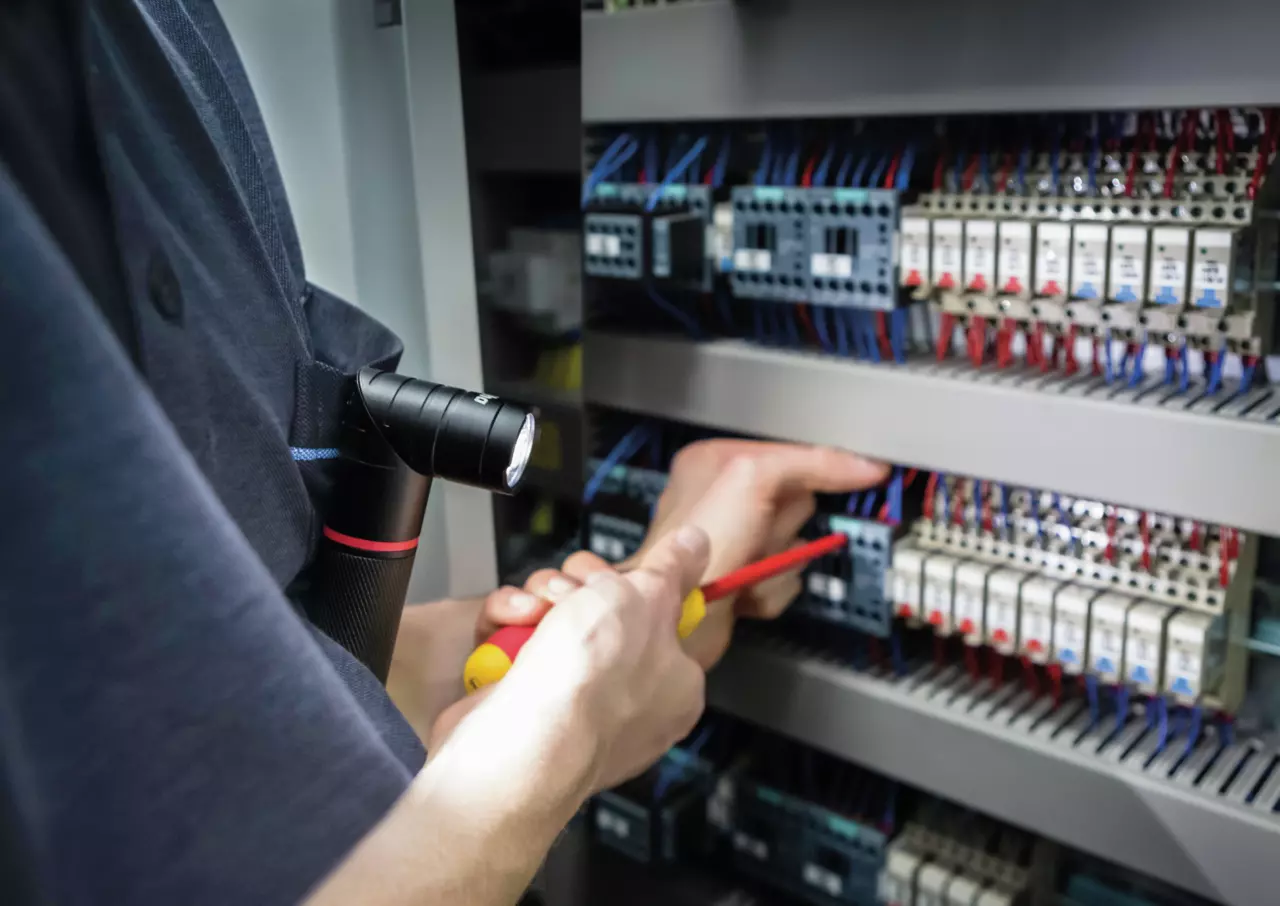 A person works on an electrical panel using a screwdriver, with a flashlight clipped to their shirt.