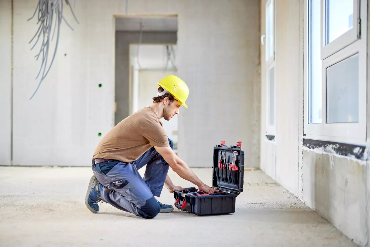 A worker in a yellow helmet kneels on the floor, reaching into a tool case in a construction site.