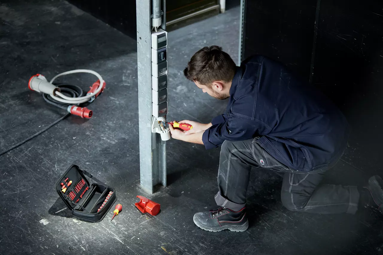 A person kneeling and using a screwdriver to work on electrical outlets on a metal beam. Tool case nearby.