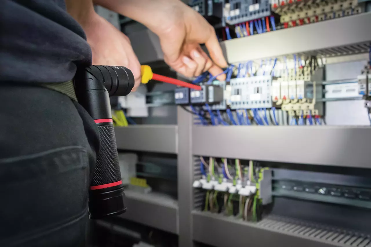 A person working with a screwdriver inside an electrical panel, a flashlight clipped to their belt.