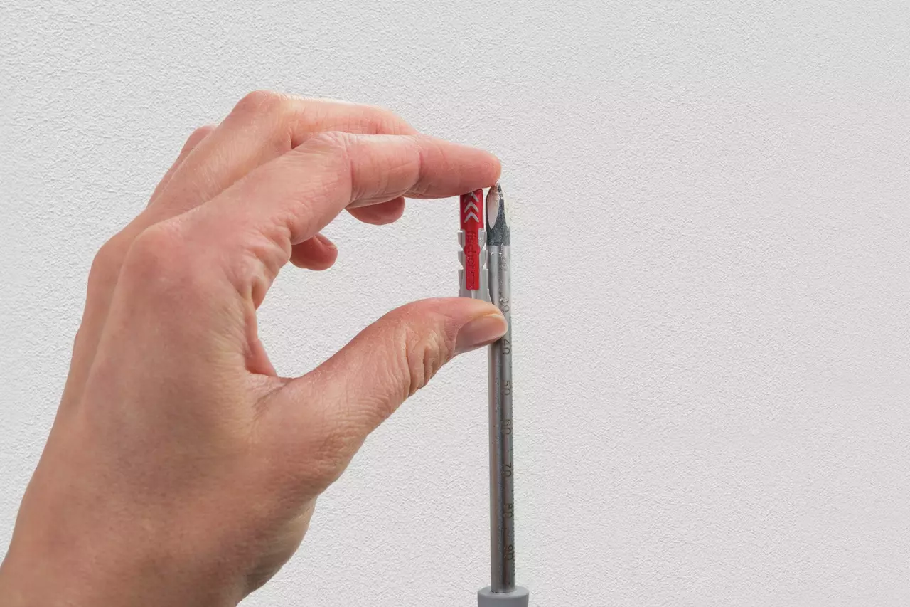 A hand holding a red wall plug next to a metal drill bit against a white background.