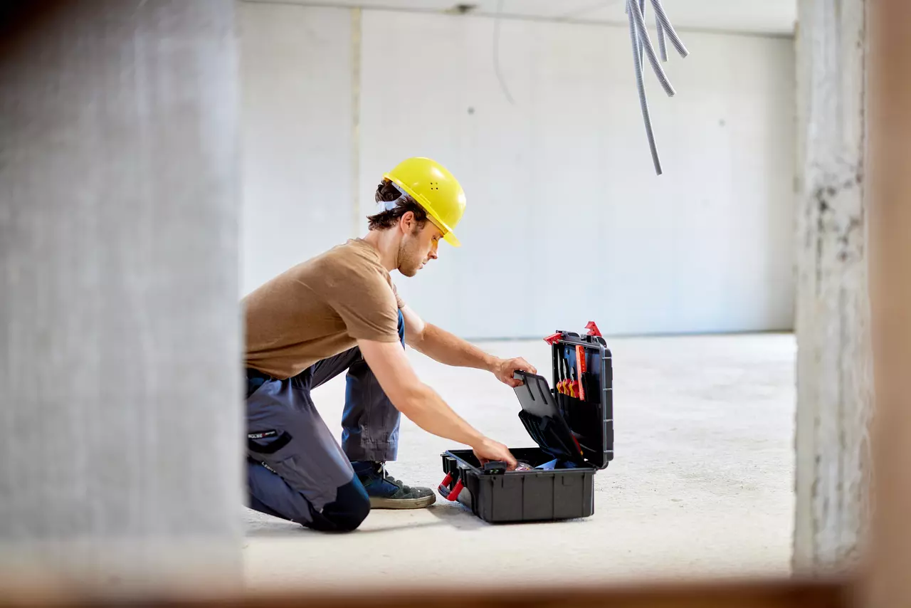 A person in a yellow hard hat kneeling and organizing tools in a toolbox in a construction area.