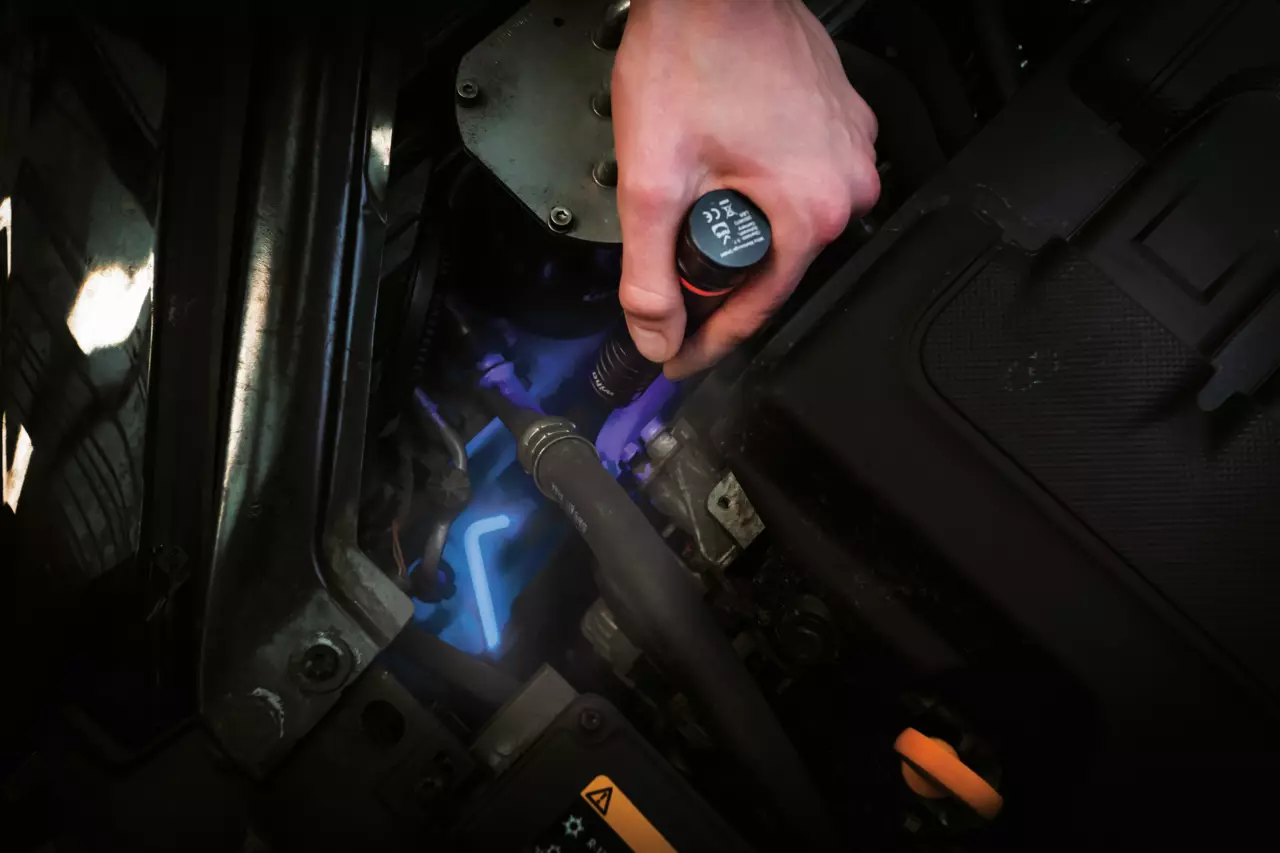 A person uses a flashlight to inspect a car engine, illuminating parts in blue light.