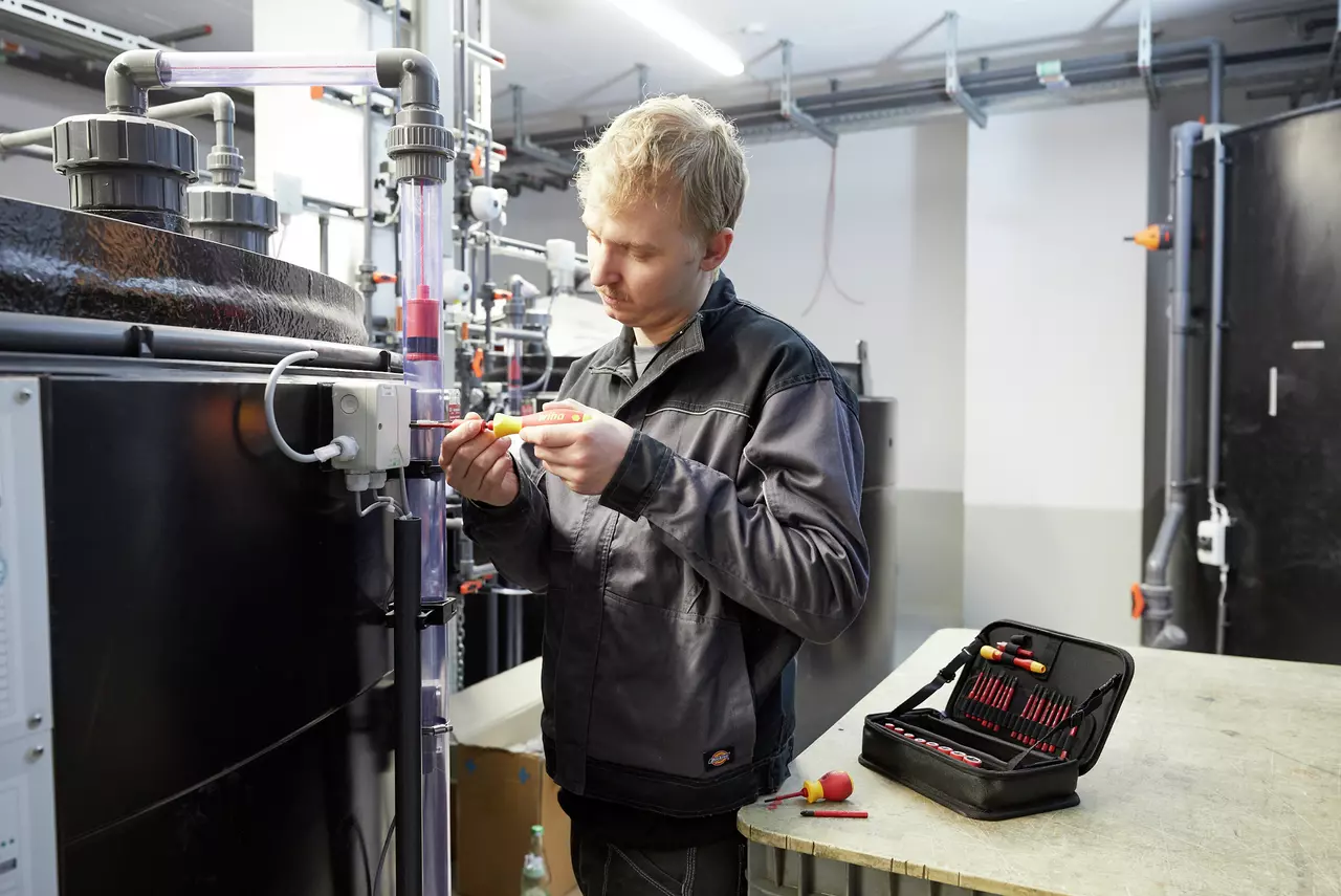 A person using a screwdriver on industrial equipment, with a tool kit on a table nearby.