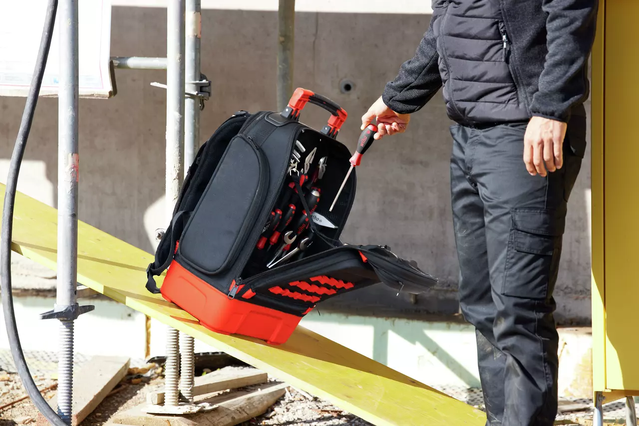 Person taking a screwdriver from a black and red tool backpack on a construction site.