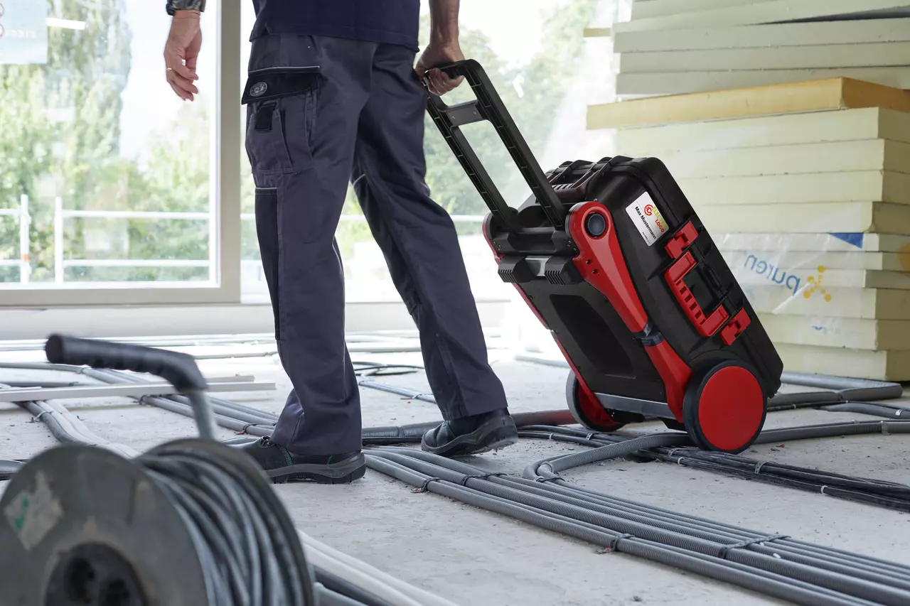 A person pulling a wheeled tool case on a construction site with cables on the floor.