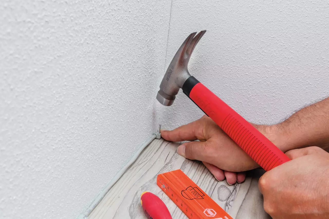 Person using a red-handled hammer to secure a cable clip on a baseboard.