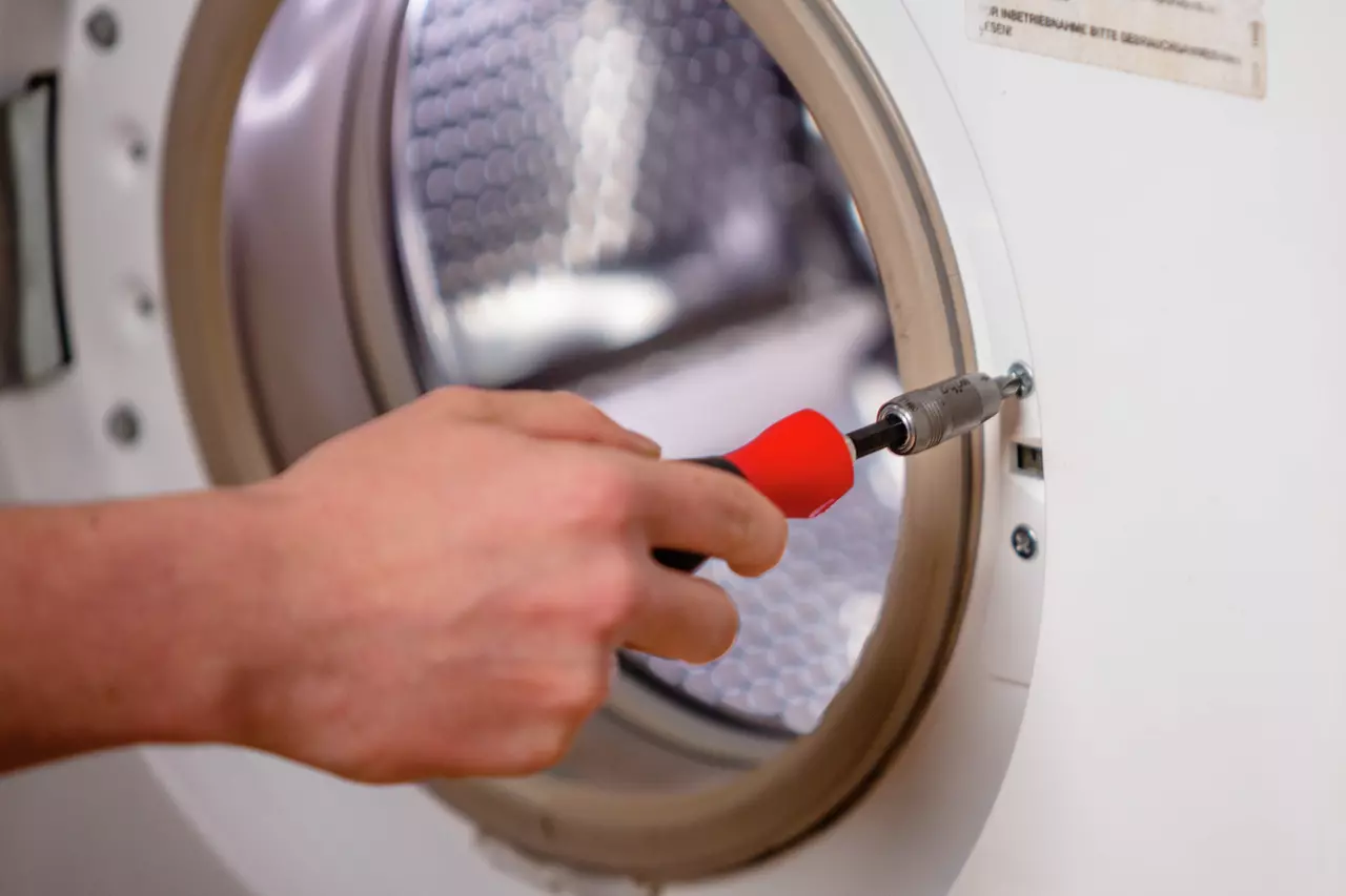 A person uses a red-handled screwdriver on a washing machine door hinge.