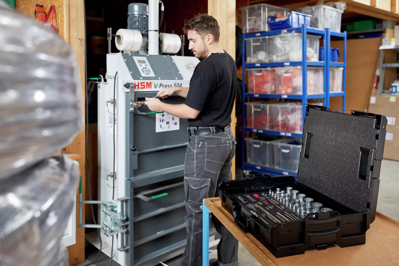A person using a wrench on a machine in a workshop, with a socket set open on a table.