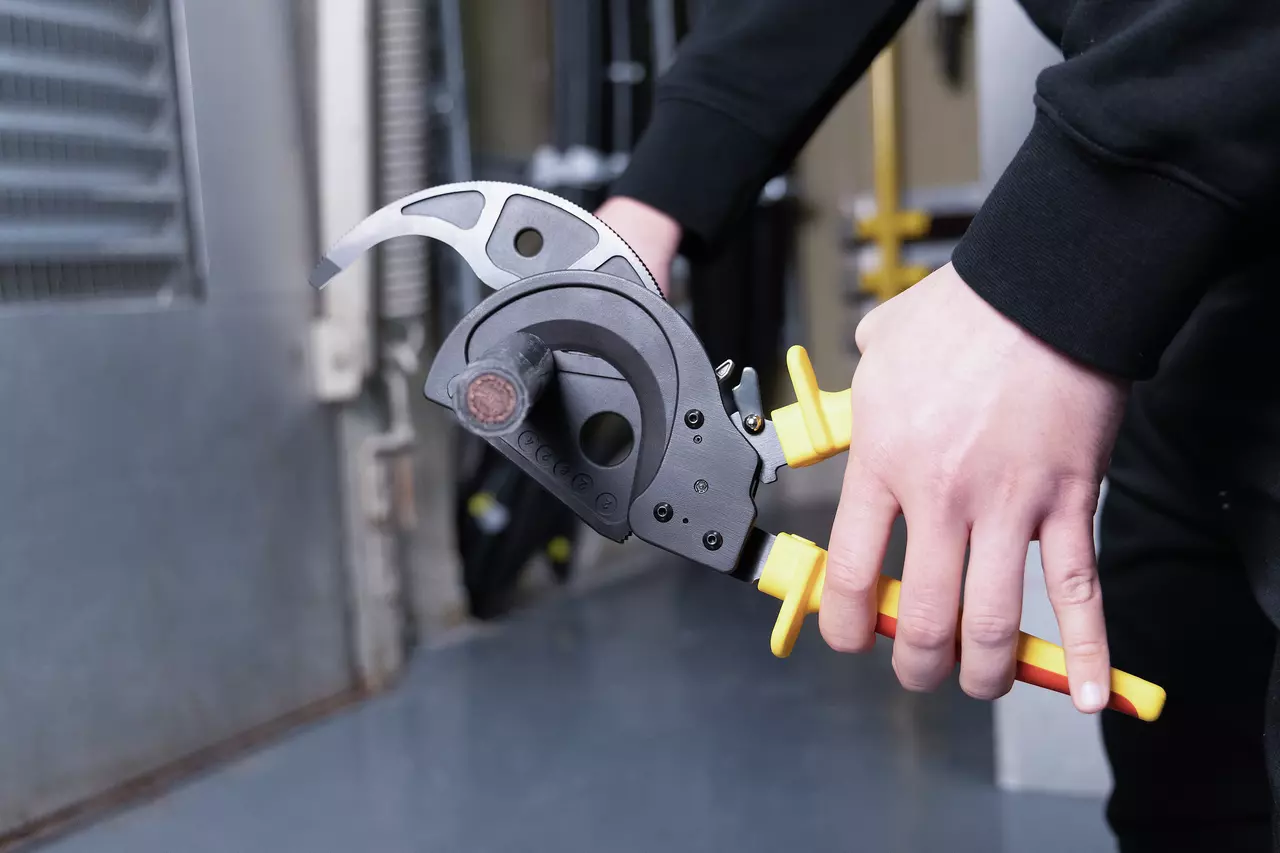 Person using a ratchet cable cutter with yellow handles to cut a thick black cable.