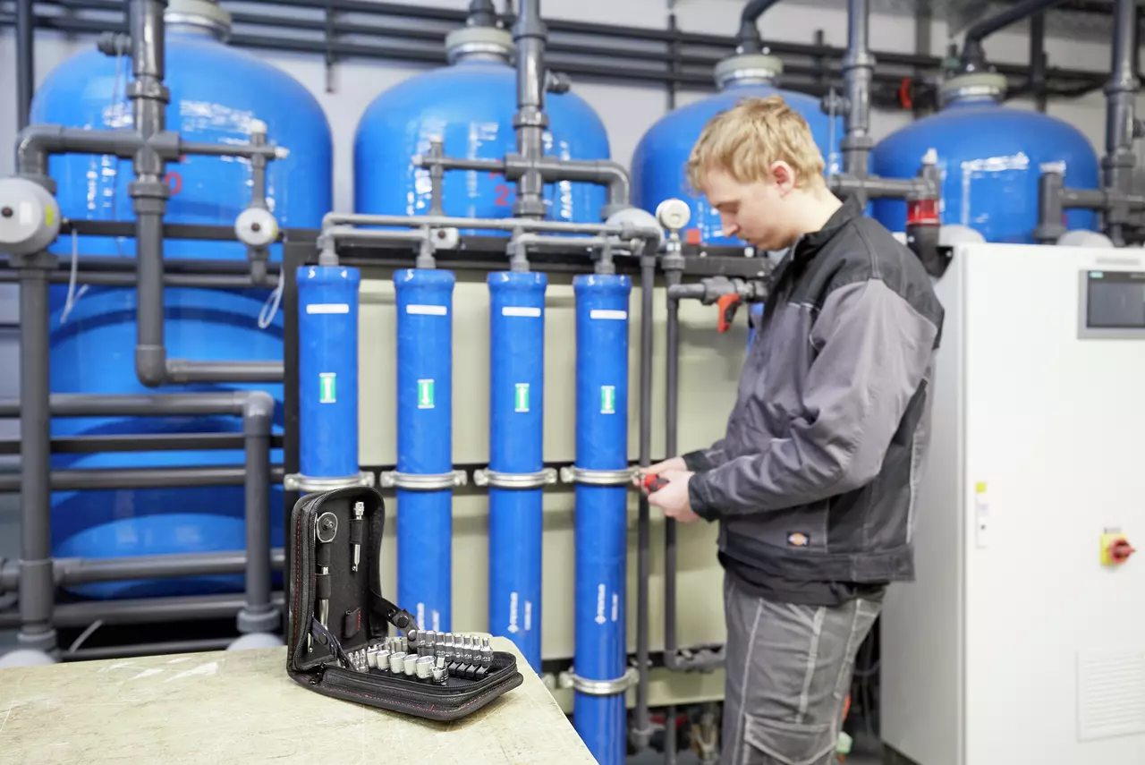 A person working in a facility with blue tanks, holding a tool. A socket set is on a table nearby.