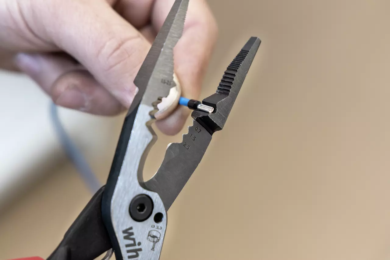Een hand gebruikt een tang om isolatie van een blauwe elektrische draad te verwijderen.