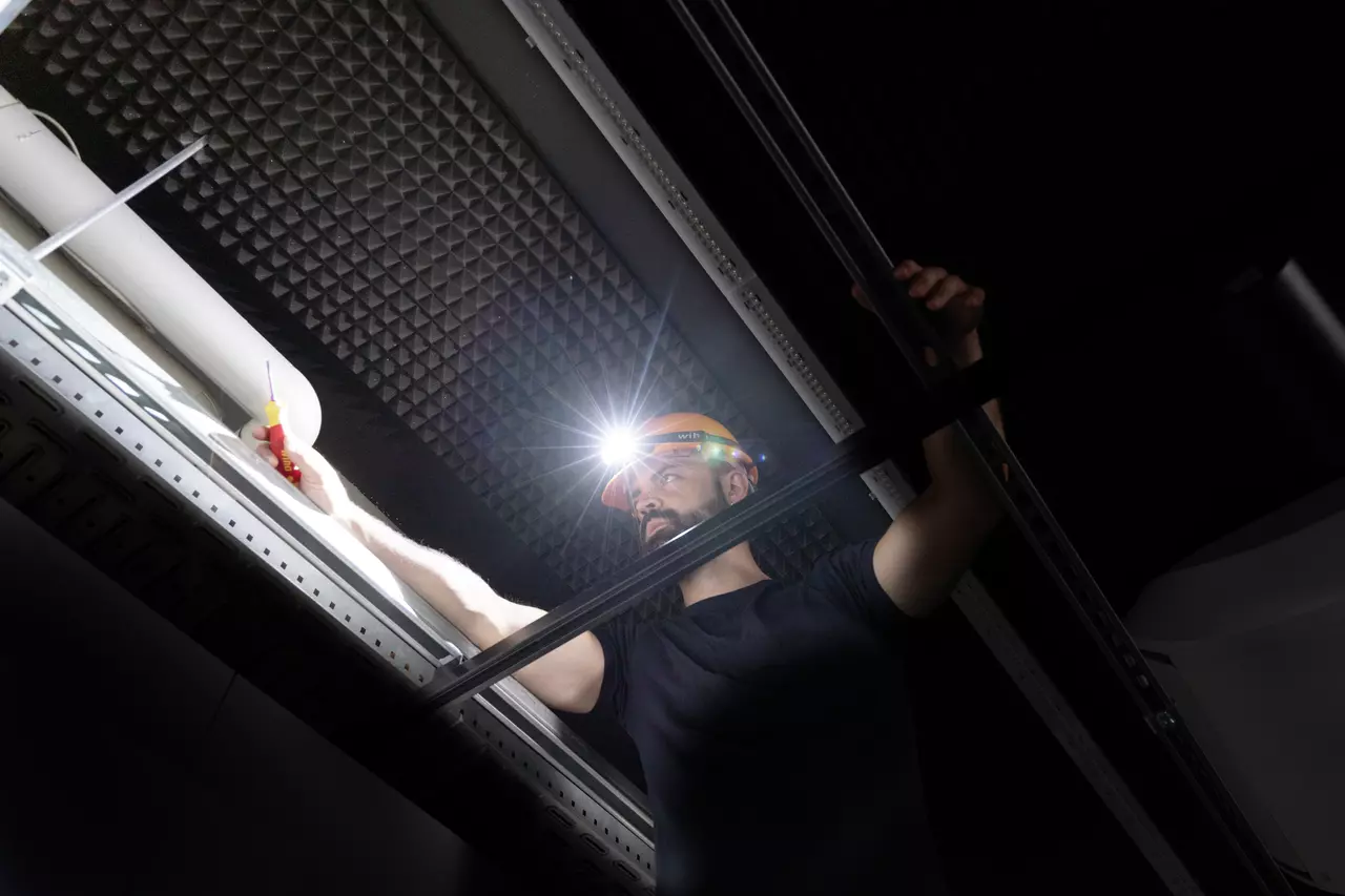 A person wearing a headlamp uses a screwdriver while standing on a stepladder in a dark room.