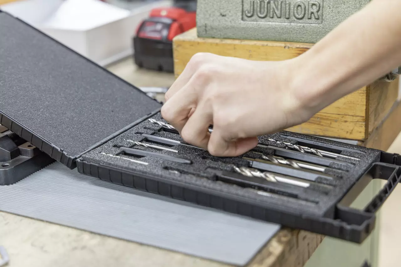 A person selecting a drill bit from a black foam-lined case containing various drill bits on a workbench.