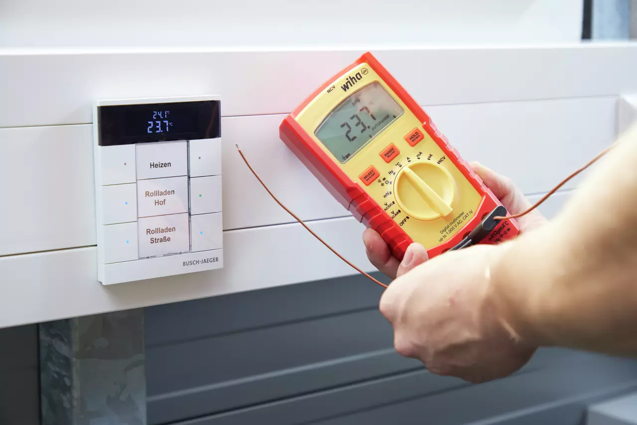 Person använder en digital multimeter som visar 23,7°C nära en kontrollpanel med temperatur- och persiennkontroller.