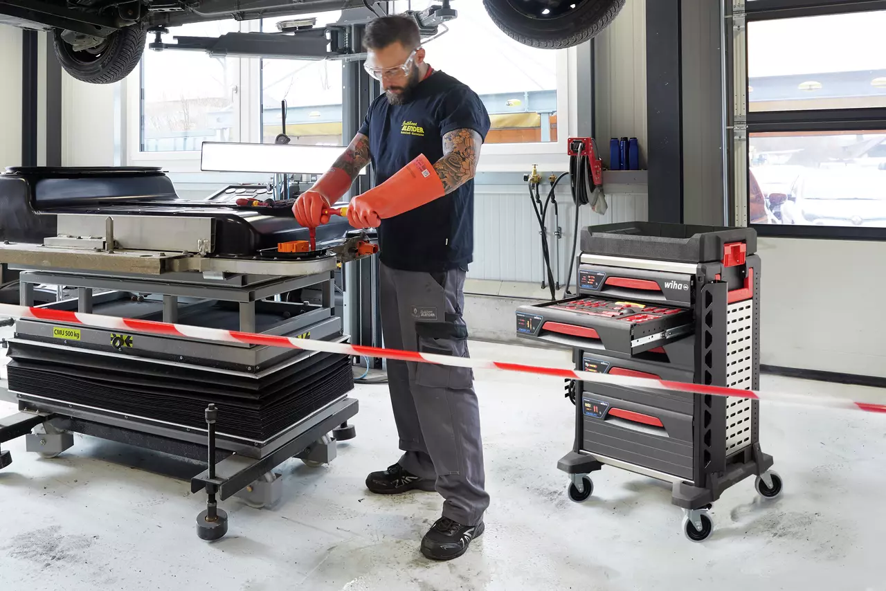 A person working on a large black component in a garage, with a tool trolley nearby.