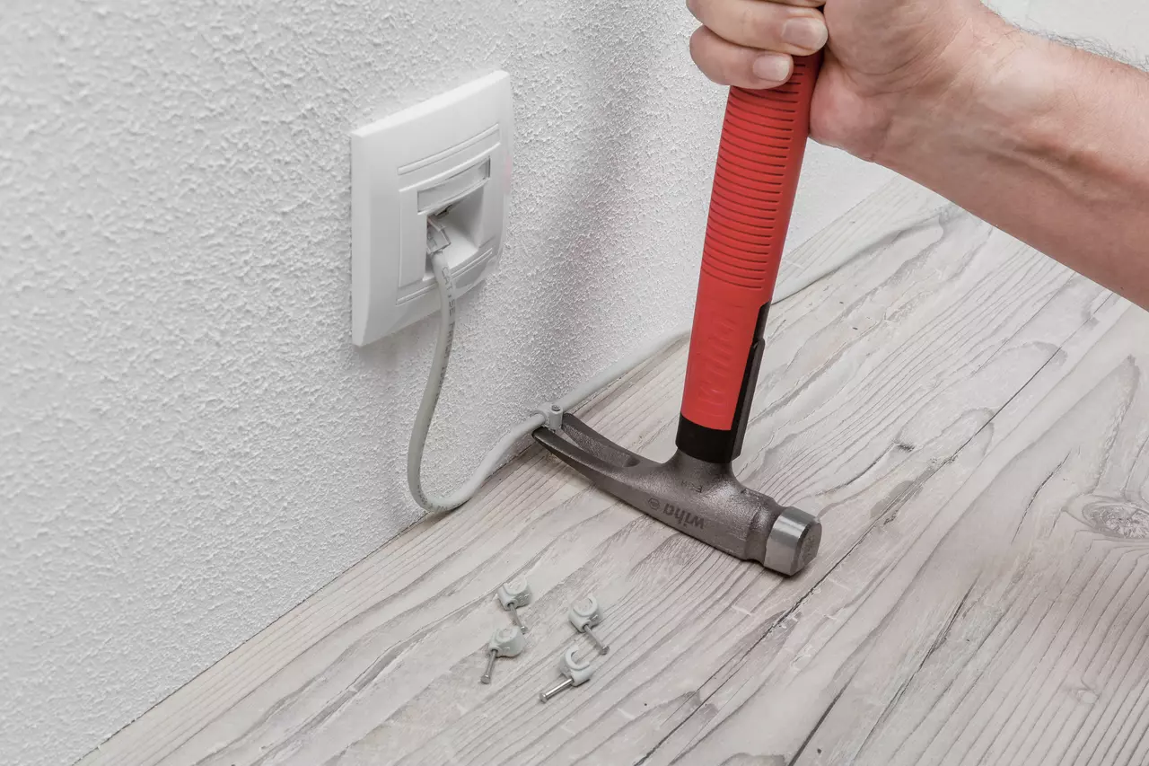 Person using a red-handled electrician's hammer to secure a cable to a wooden floor with clips.