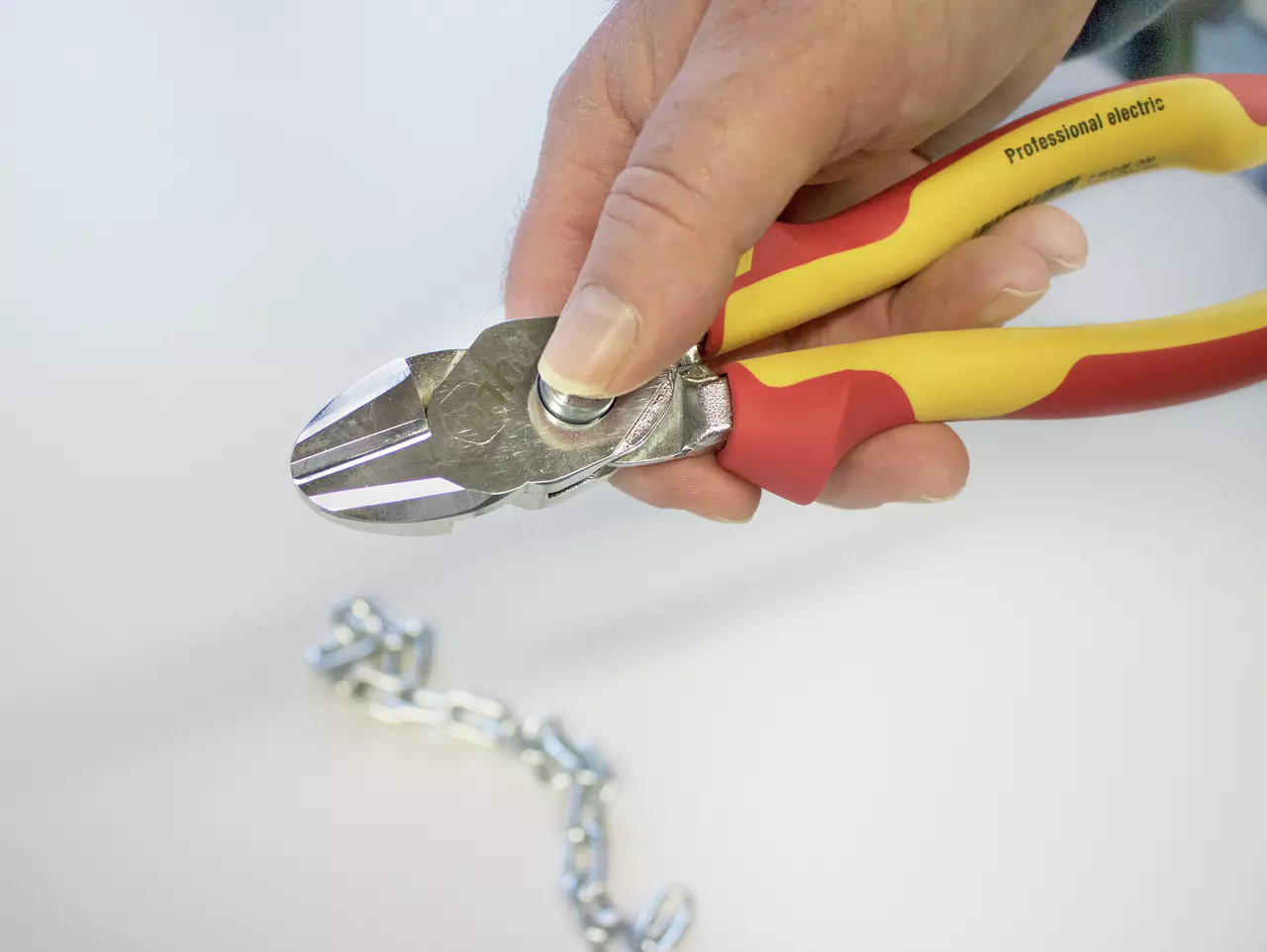 A hand using insulated pliers to cut a metal chain on a white surface.