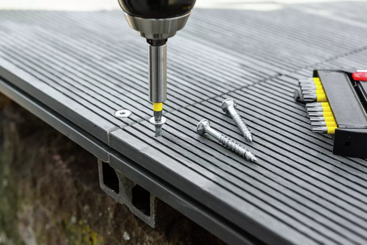 Electric screwdriver driving a screw into a grooved decking board, with additional screws and bits nearby.