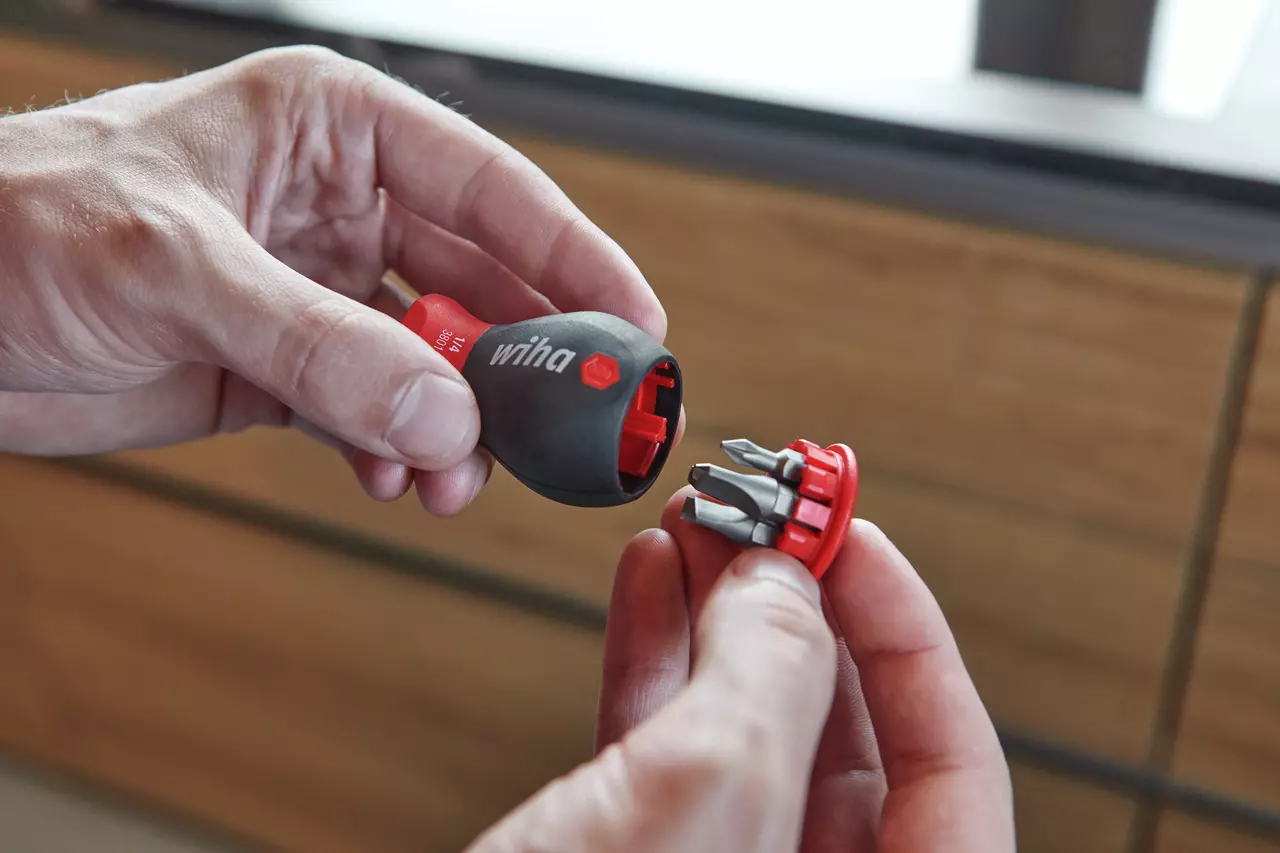 Two hands holding a screwdriver handle and a bit holder with multiple bits. The handle is black and red.