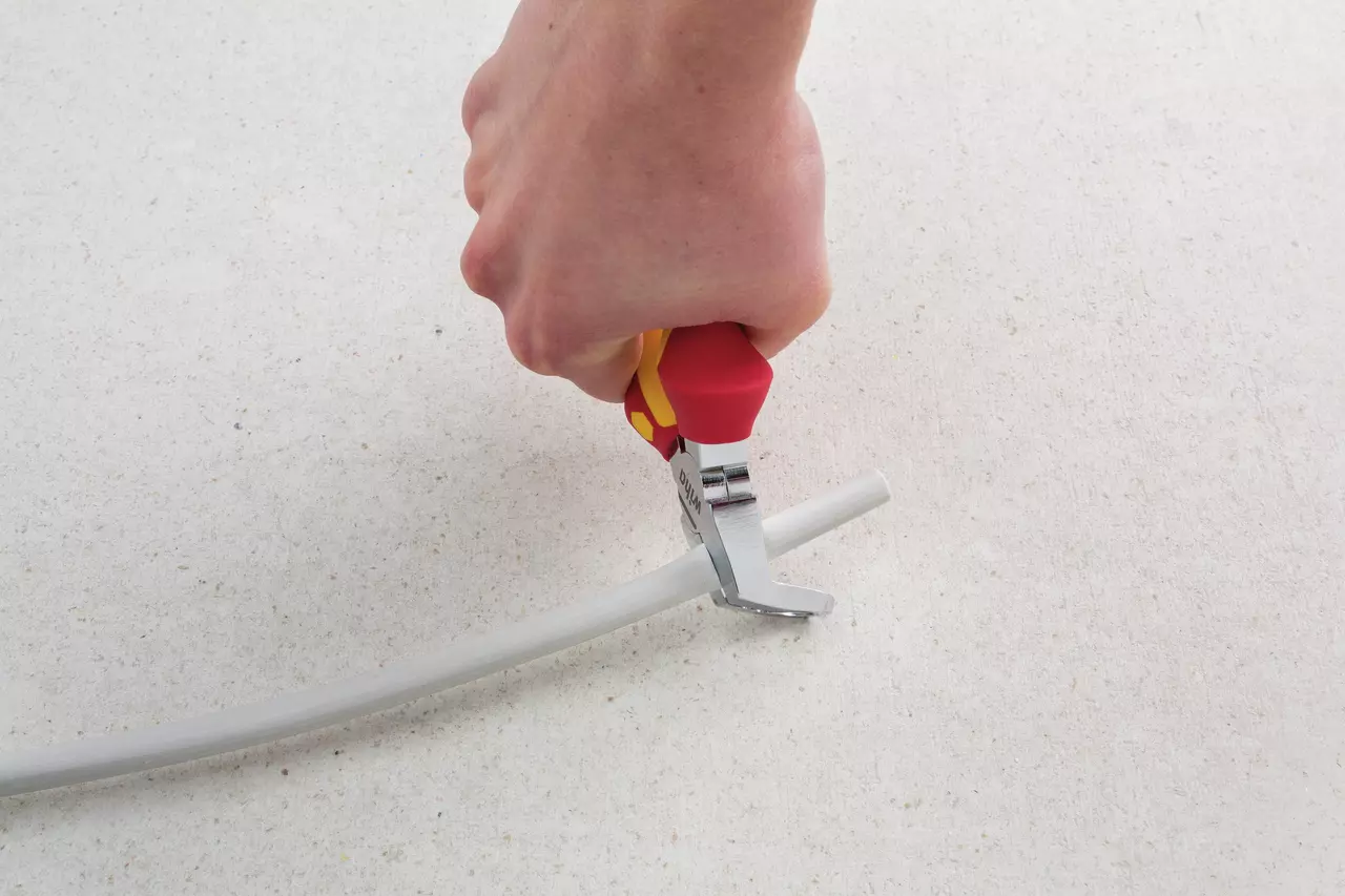 Hand using red-handled tool to cut a gray cable on a light surface.