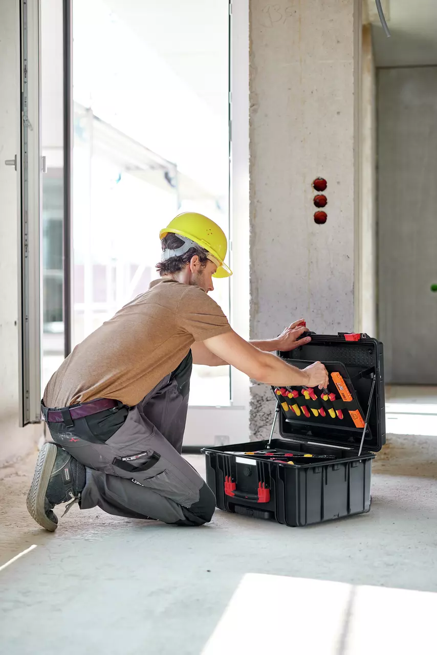 A person in a yellow hard hat kneels, organizing tools in a black toolbox on a construction site.