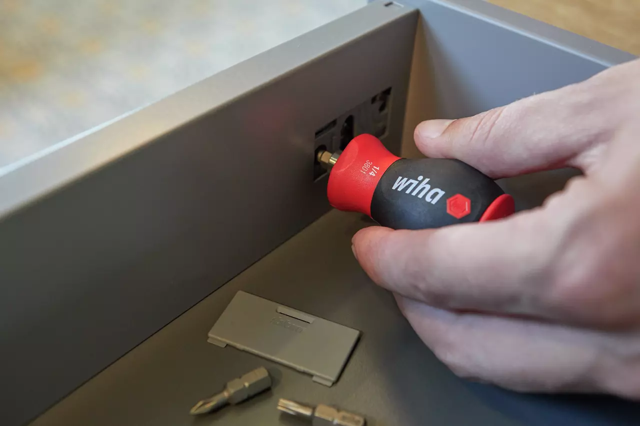 A person uses a red and black screwdriver on a drawer. Bits are placed nearby.