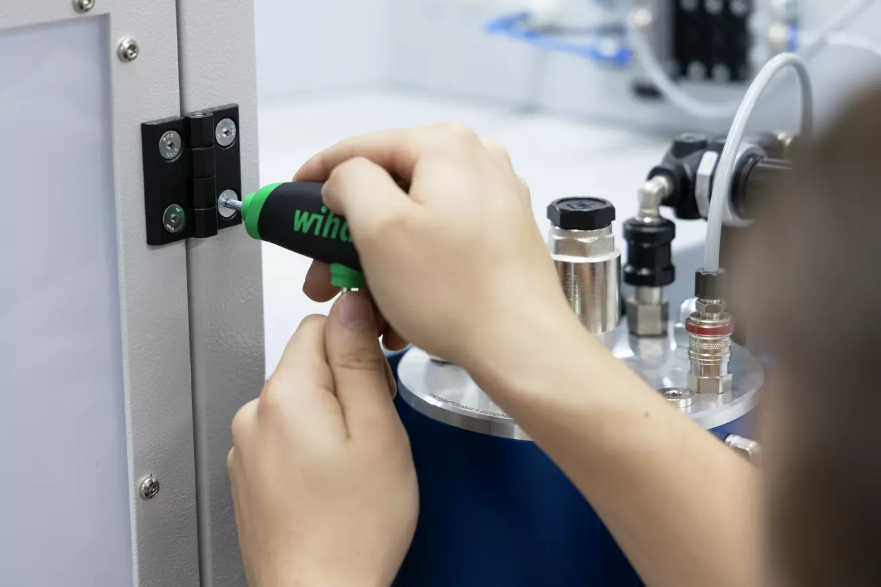 Person using a green-handled tool on a hinge attached to a metal panel.