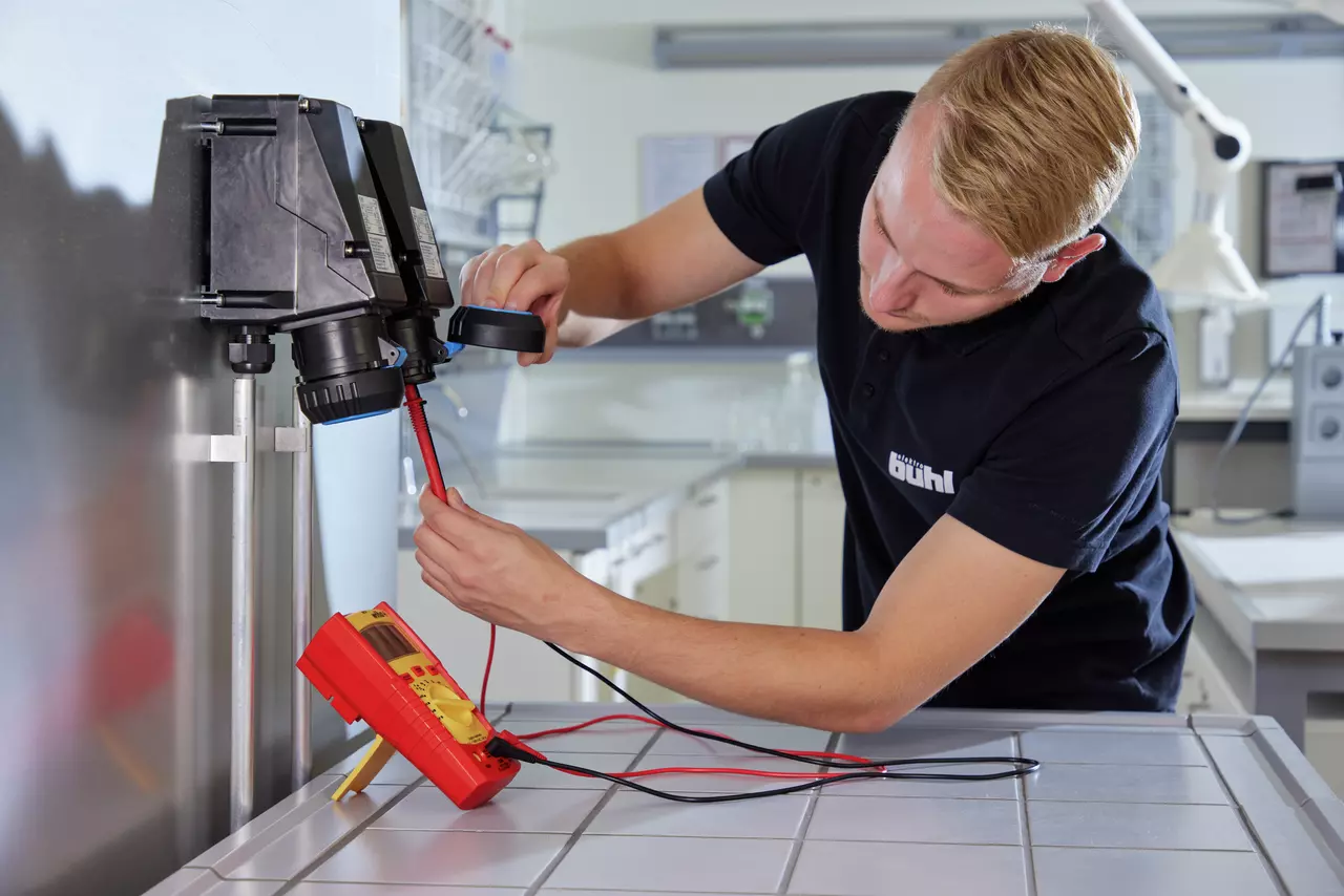 A person using a digital multimeter with red probes on a black device in a lab setting.