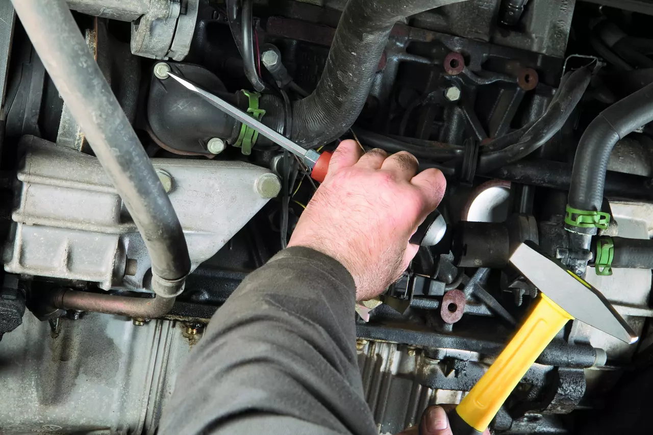 A person using a screwdriver on a car engine, with a hammer in the other hand.