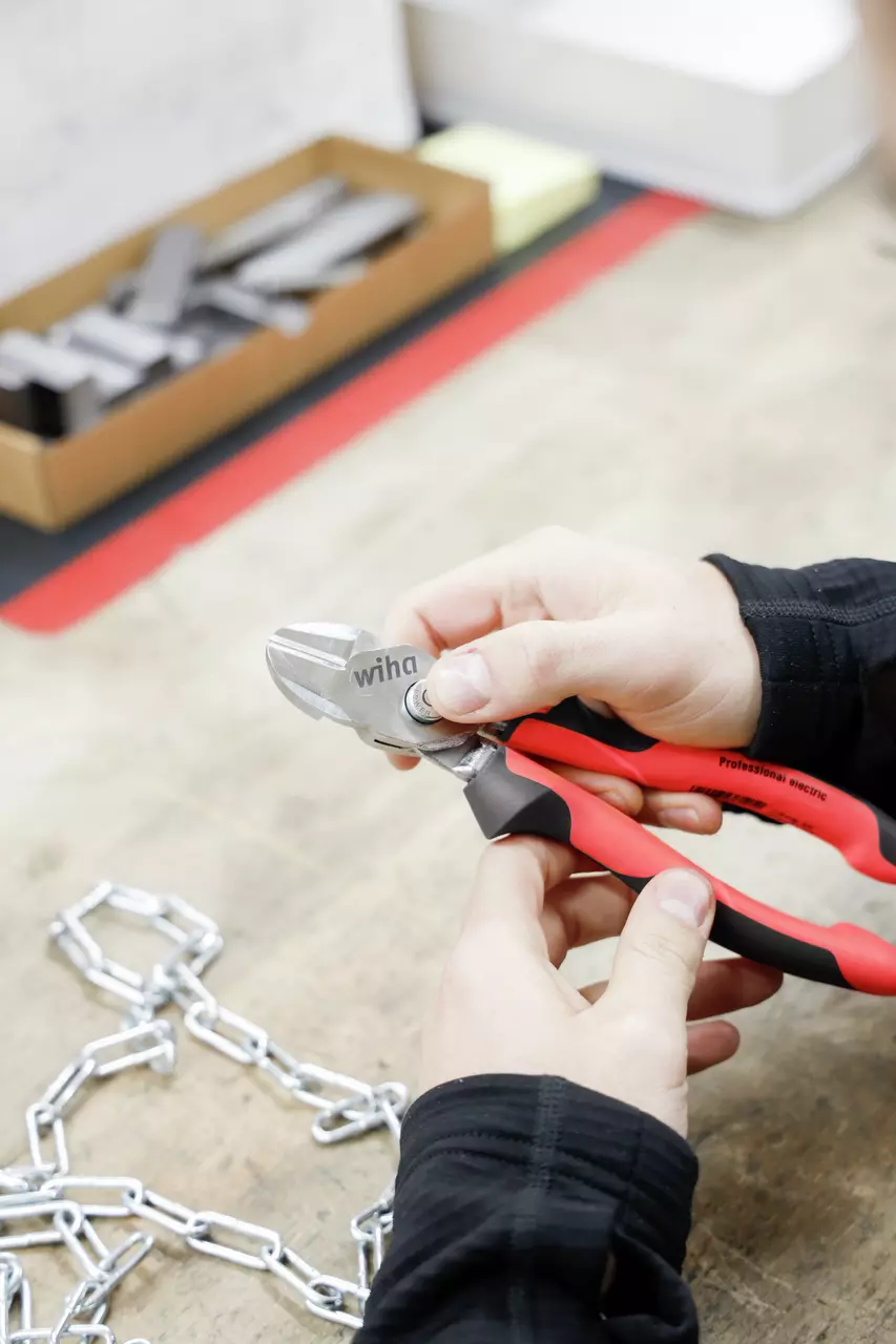 Hands holding red-handled pliers cutting a metal chain on a workbench.