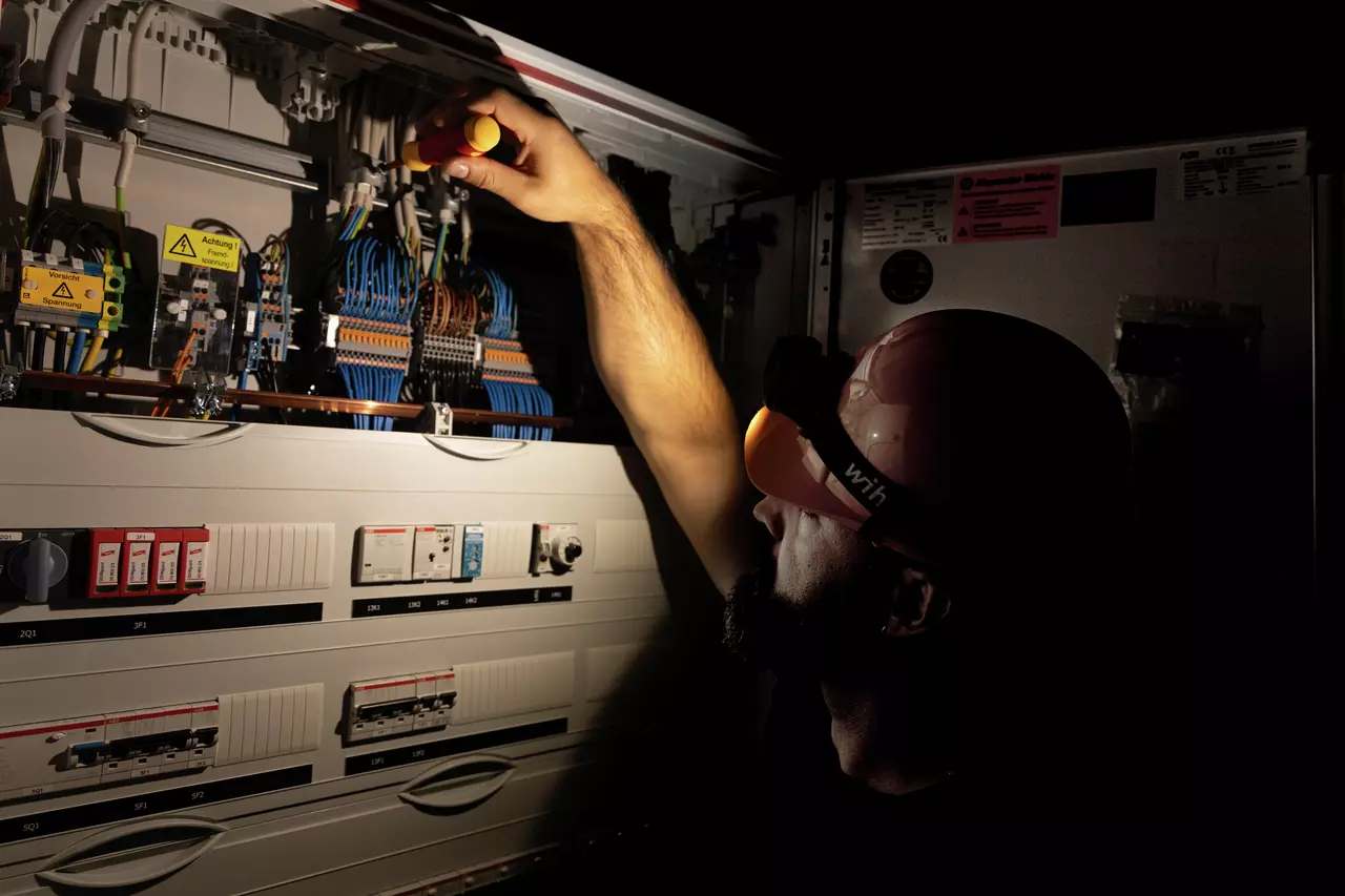 A person wearing a helmet works on an electrical cabinet with a screwdriver, surrounded by wires and switches.