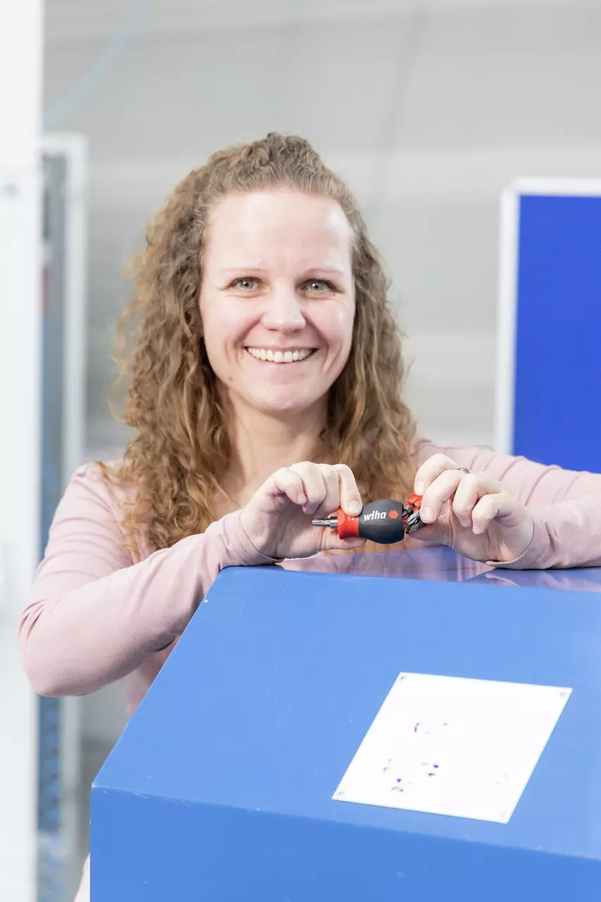 A woman holding a black and red screwdriver with bit storage, standing by a blue surface.