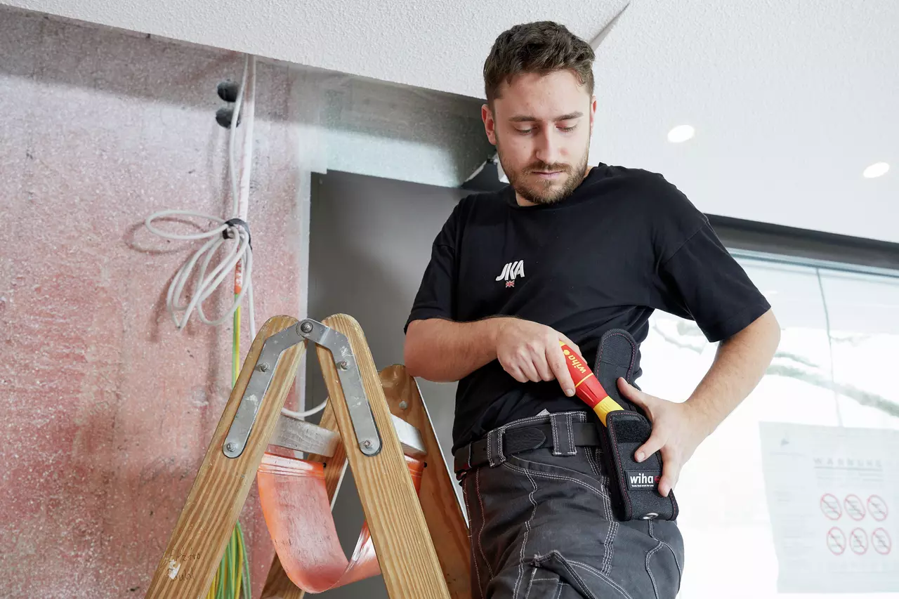 A person on a ladder holds a red and yellow electrical tool in a holster. Wires are visible in the background.