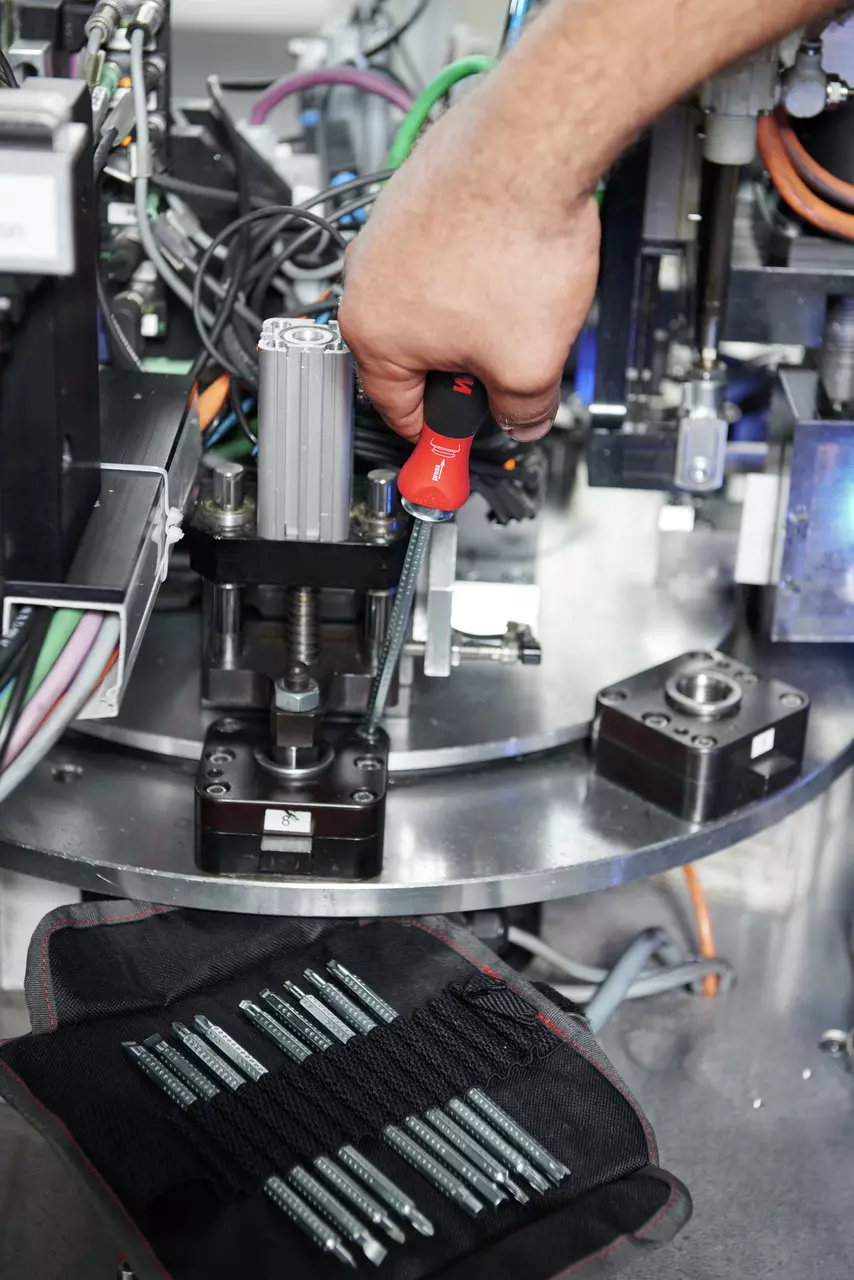 Person using a red-handled screwdriver on machinery with tool bits nearby.