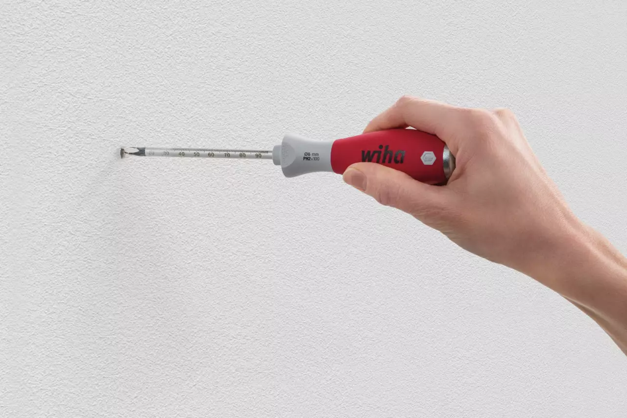 Hand holding a red tool with a measurement scale, inserted into a wall hole.