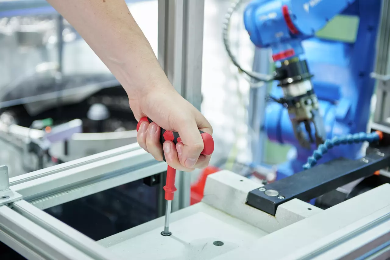 A person uses a red-handled screwdriver on a metal frame in an industrial setting with a blue robotic arm nearby.