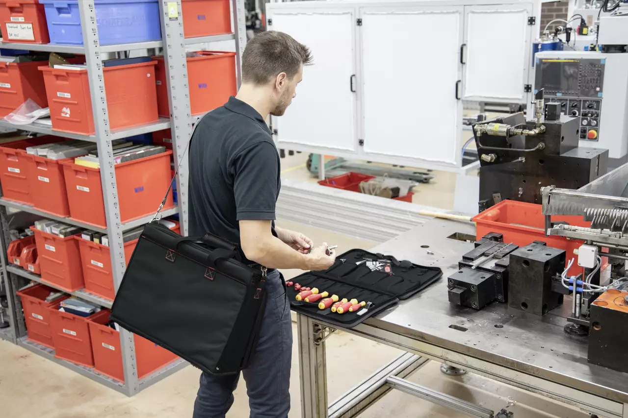 A person in a workshop with a tool set open on a table, surrounded by machinery and red storage boxes.
