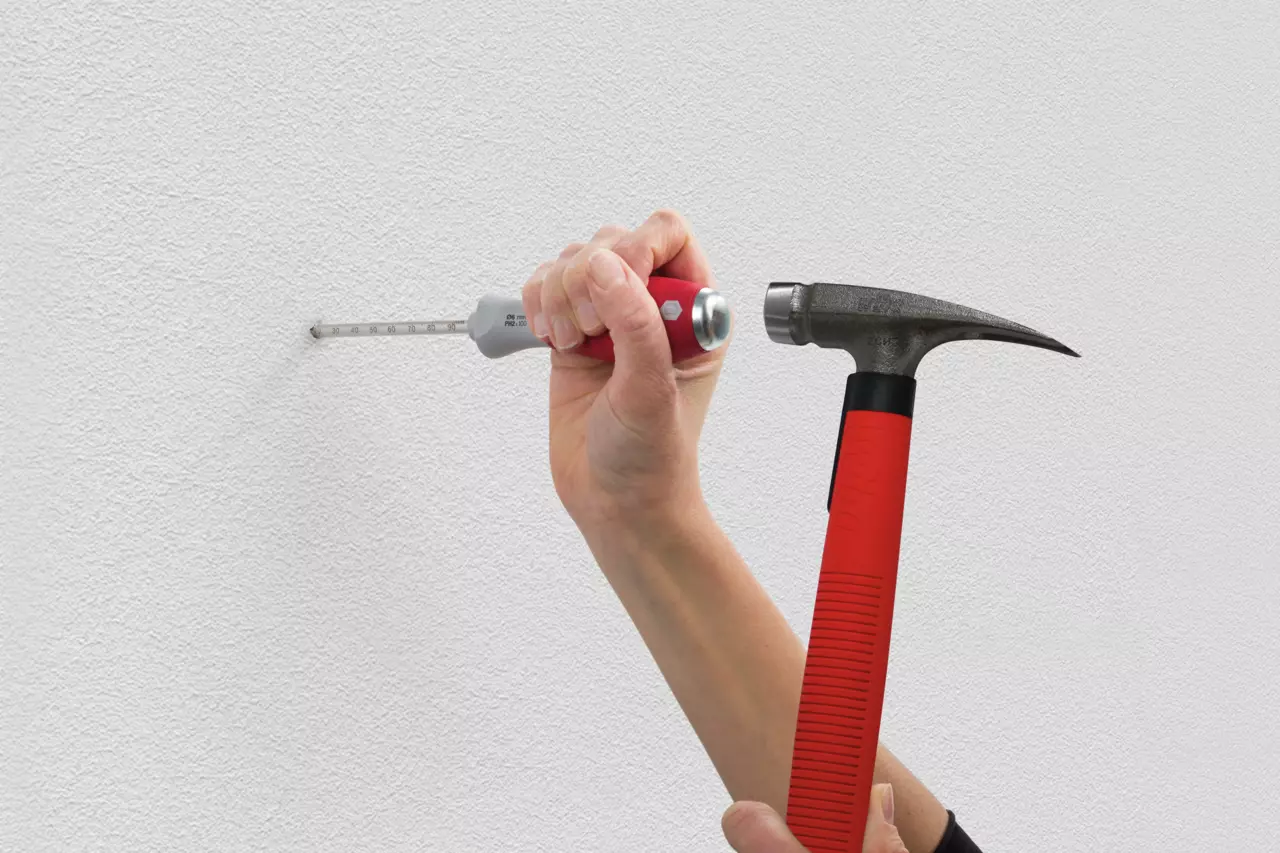 Hand using a hammer and tool to measure a hole in a white wall