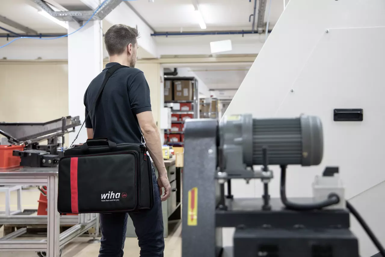 A person in a workshop carrying a black tool bag with red accents.