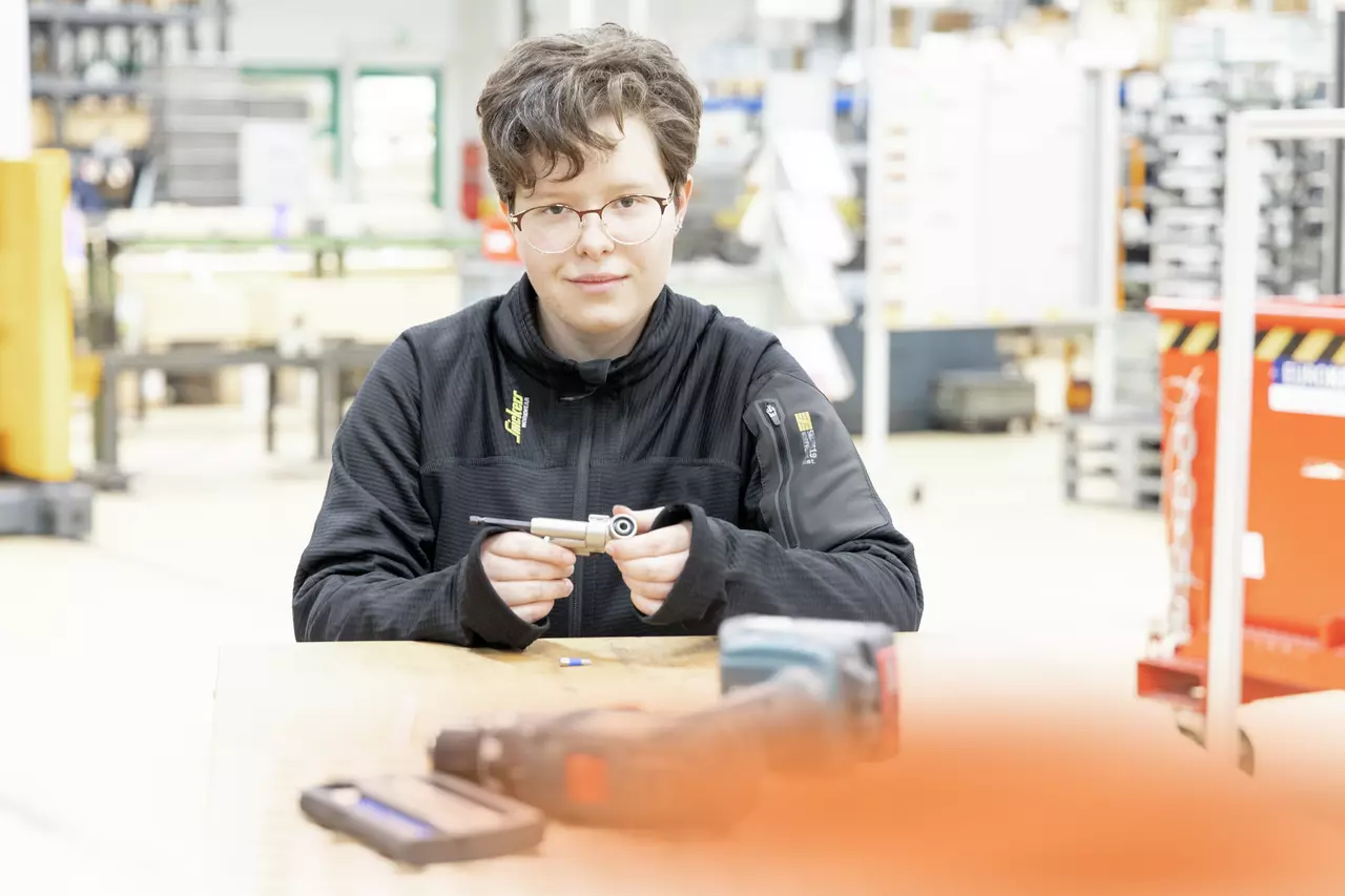 A person with glasses holds a tool in a workshop.