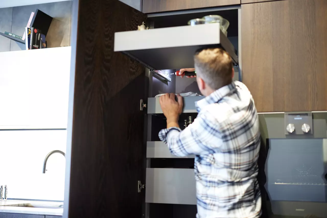 A person uses a screwdriver inside a kitchen cabinet.