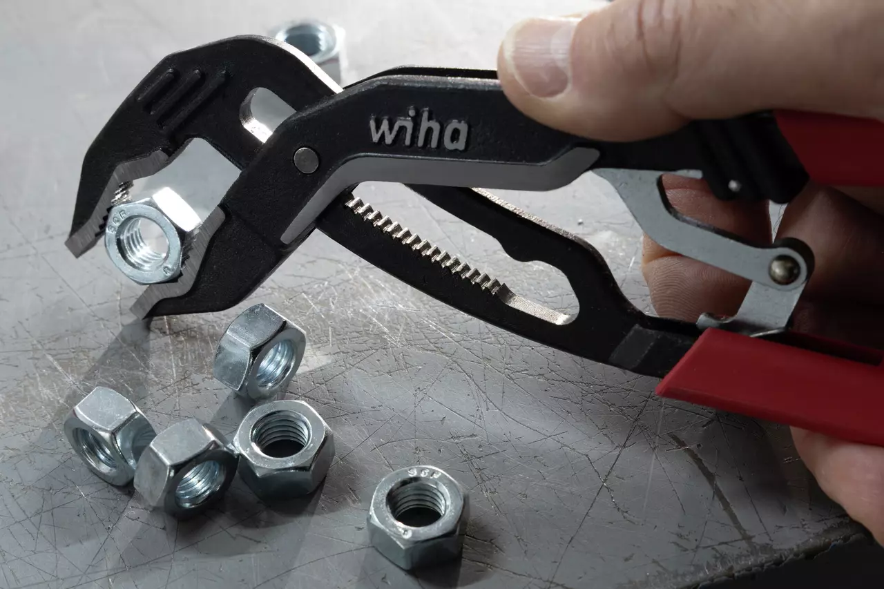 Adjustable pliers gripping a metal nut, surrounded by loose nuts on a scratched metal surface.